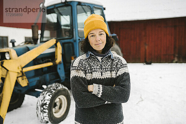 Porträt einer reifen Frau  die im Winter mit verschränkten Armen vor einem Traktor steht