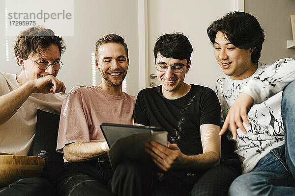 Lächelnder Mann  der ein digitales Tablet benutzt  während er sich mit männlichen Freunden im Wohnzimmer austauscht