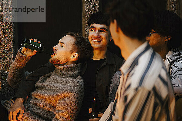 Junger Mann trinkt Bier beim Chillen mit männlichen Freunden auf dem Balkon