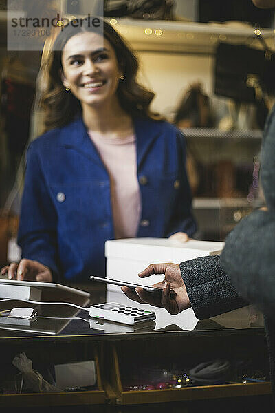 Lächelnder weiblicher Besitzer  der den Kunden beim kontaktlosen Bezahlen im Geschäft betrachtet