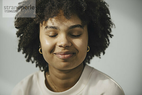 Lächelnde junge Frau mit geschlossenen Augen gegen weiße Wand