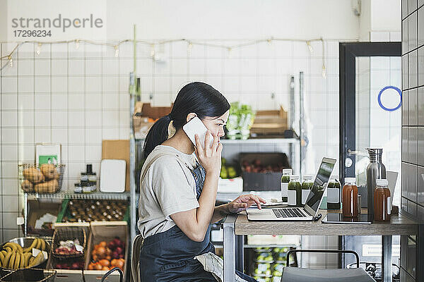Weibliche Unternehmerin im Gespräch über Smartphone während der Arbeit am Laptop im Geschäft