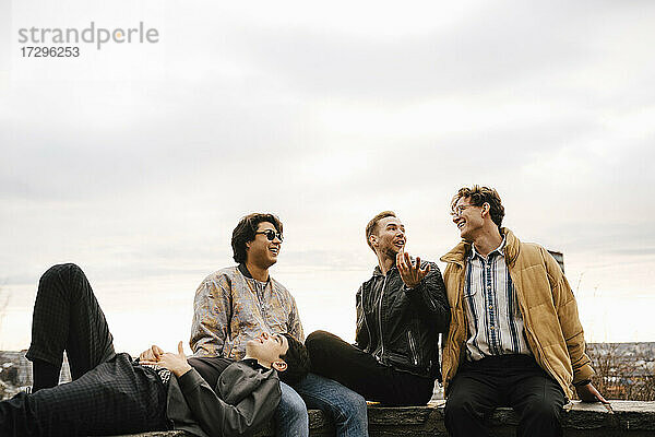 Junge männliche Freunde verbringen ihre Freizeit  während sie auf einer Stützmauer gegen den Himmel sitzen