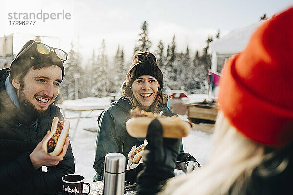 Glückliche Freunde essen Hot Dog beim Sitzen im Skigebiet im Winter