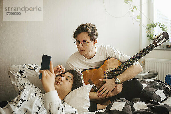 Mann mit Gitarre guckt  während männlicher Freund mit Smartphone im Schlafzimmer