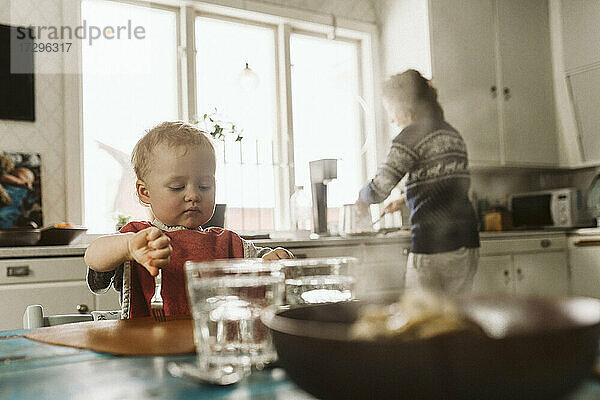 Mädchen sitzt am Esstisch  während die Mutter das Essen in der Küche vorbereitet