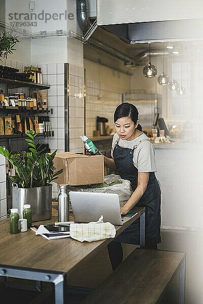 Weiblicher Besitzer arbeitet am Laptop  während er ein Lebensmittelpaket im Laden hält