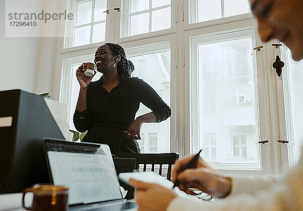 Lächelnde Geschäftsfrau mit Kaffeetasse am Fenster stehend  während ein männlicher Kollege im Home Office schreibt