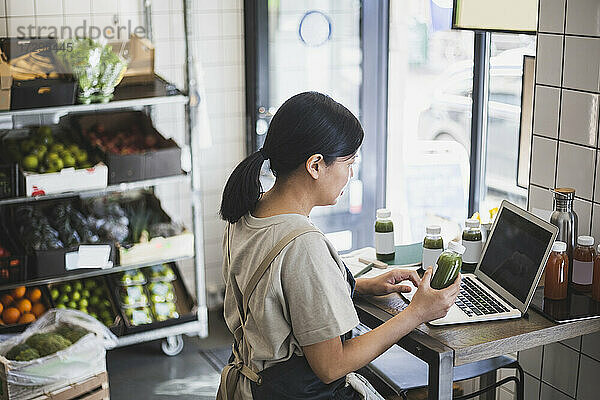 Asiatische Unternehmerin mit Laptop  während sie eine Saftflasche im Laden hält