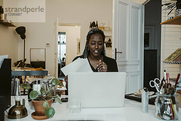 Junge Geschäftsfrau mit Dokument diskutieren auf Video-Anruf durch Laptop am Schreibtisch im Home-Office