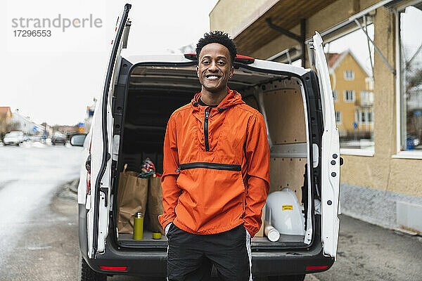 Porträt eines lächelnden Servicemitarbeiters  der mit den Händen in den Taschen vor einem Lieferwagen steht