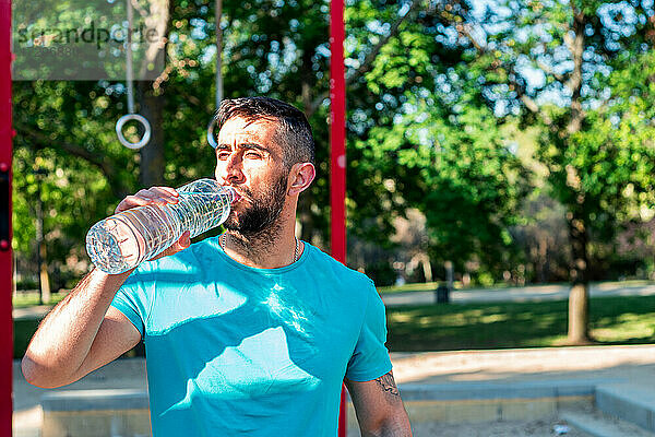 Bärtiger brünetter Mann trinkt Wasser nach dem Training in einem Park. Outdoor-Fitness-Konzept.