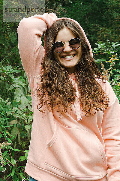 Eine junge Frau mit rosa Kapuzenpulli und Sonnenbrille  mit lockigem Haar  lächelt.