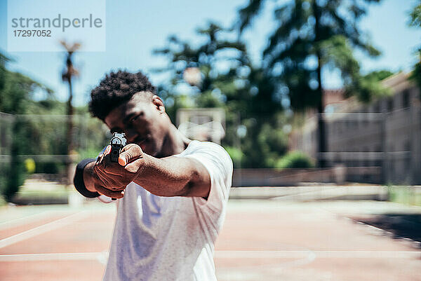 Schwarzer afroamerikanischer Junge auf einem Basketballplatz  der mit einer Waffe auf die Kamera zielt. Fokus auf die Waffe.