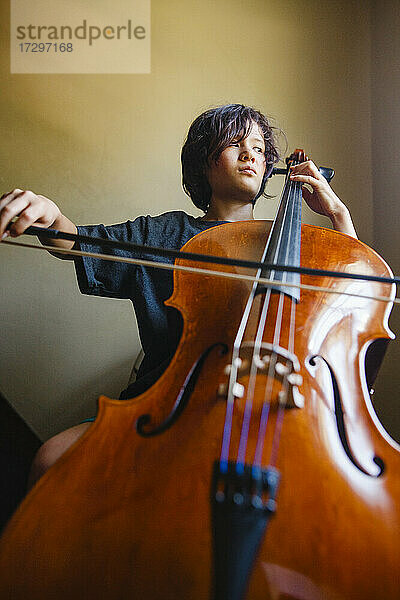 Ein Junge mit ernster Miene spielt Cello und schaut aus dem Fenster