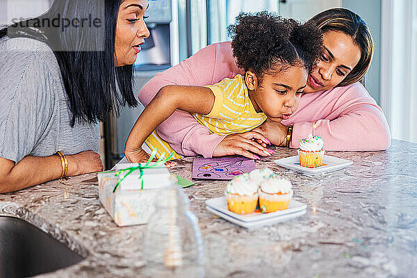 Mädchen bläst eine Kerze auf einem Kuchen aus  während sie mit Mutter und Großmutter zu Hause Geburtstag feiert