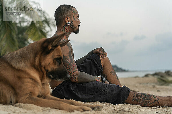 Asiatischer Mann sitzt am Strand mit einem Hund unter Palmen