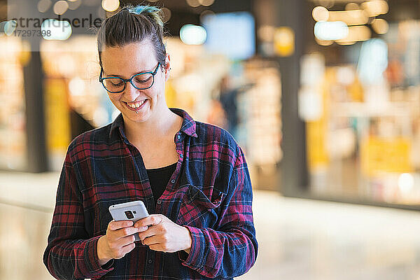 Spanische Frau lächelt und schreibt eine SMS auf ihrem Smartphone  während sie in einem Einkaufszentrum einkauft