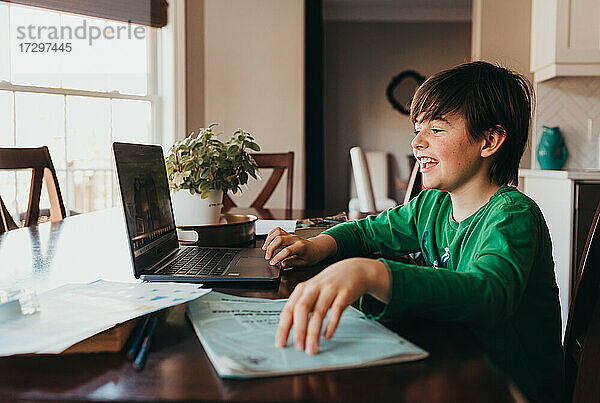 Glücklicher Junge  der am Computer am Küchentisch online Schularbeiten erledigt.