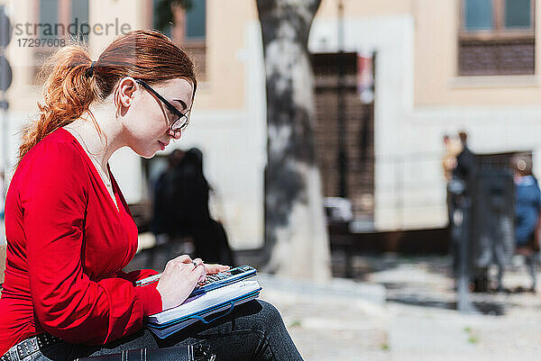 Junge rothaarige Frau mit Brille sitzt auf einer Bank  benutzt einen Taschenrechner und schreibt etwas in ihr Notizbuch. Bekleidet mit einer roten Bluse.