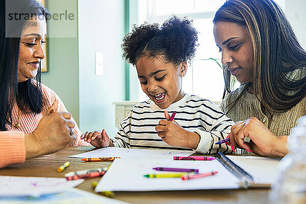 Mutter und Großmutter mit fröhlichem Mädchen  das mit Buntstiften auf Papier zu Hause zeichnet