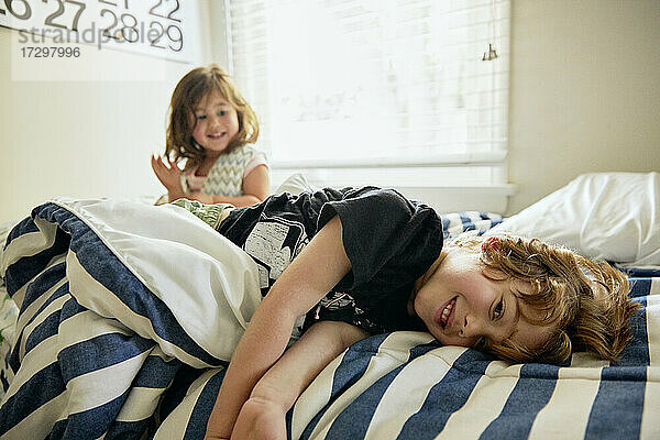 Porträt eines lächelnden Jungen  der neben seiner Schwester auf dem Bett im heimischen Schlafzimmer liegt