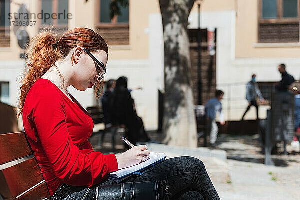 Junge rothaarige Frau mit Brille und roter Bluse sitzt auf einer Bank und schreibt etwas in ihr Notizbuch.