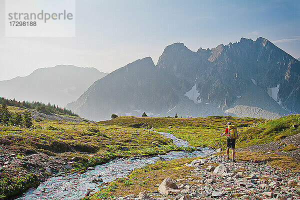 Wanderer mit Rucksack wandert neben einem ruhigen Bach auf einer Bergwiese.