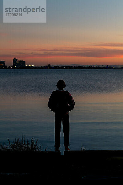 Silhouette einer jungen Person  die bei Sonnenuntergang über die Bucht auf die Stadt blickt