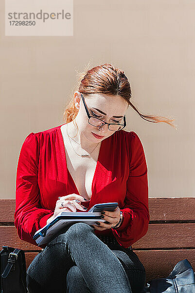Junge rothaarige Frau mit Brille sitzt auf einer Bank  benutzt einen Taschenrechner und schreibt etwas in ihr Notizbuch. Bekleidet mit einer roten Bluse.