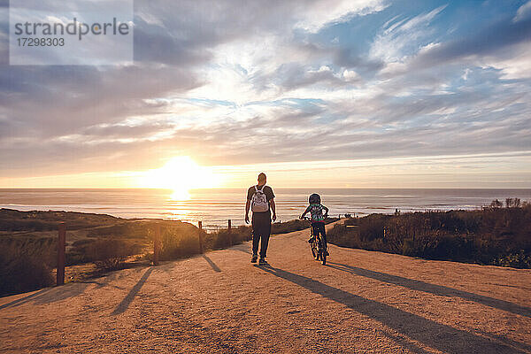 Vater beobachtet seinen Sohn beim Radfahren auf einem Küstenweg bei Sonnenuntergang.
