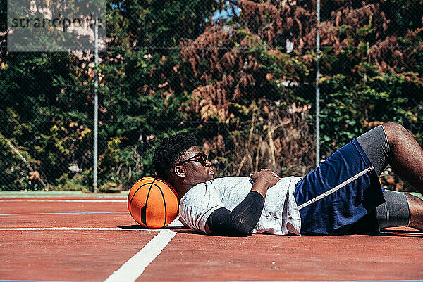 Schwarzer Afro-Typ  der auf seinem Ball auf dem Basketballplatz liegt  während er sein Handy benutzt. Konzept der Technologie und Sport.