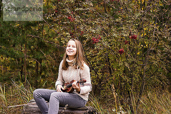 Ein hübsches Teenager-Mädchen mit langen blonden Haaren sitzt auf einem Baumstamm im Park und hält eine Geige in den Händen  schaut glücklich