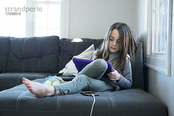 Kleines Mädchen sitzt auf der Couch und benutzt ein Tablet.
