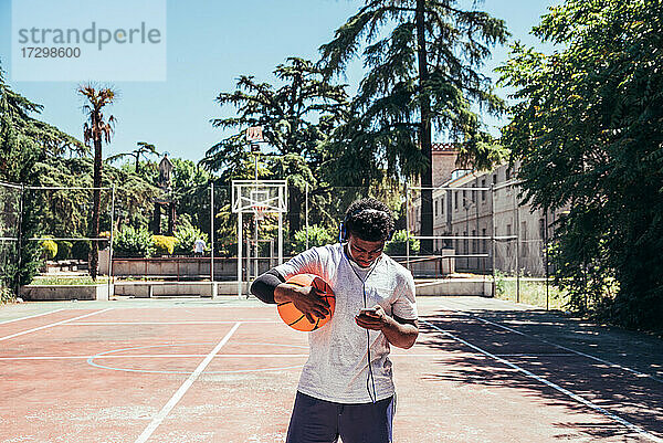 Schwarzer afroamerikanischer Junge  der mit Kopfhörern und seinem Mobiltelefon Musik hört und auf einem städtischen Platz Basketball spielt.