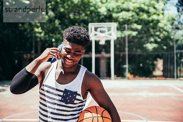 Porträt eines schwarzen afroamerikanischen Jungen  der auf einem städtischen Basketballplatz mit seinem Mobiltelefon spricht.