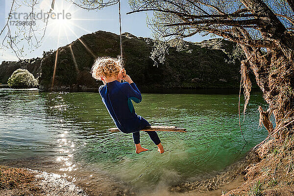 Blondes Kind auf einer Schaukel über einem Fluss in Neuseeland an einem sonnigen Tag