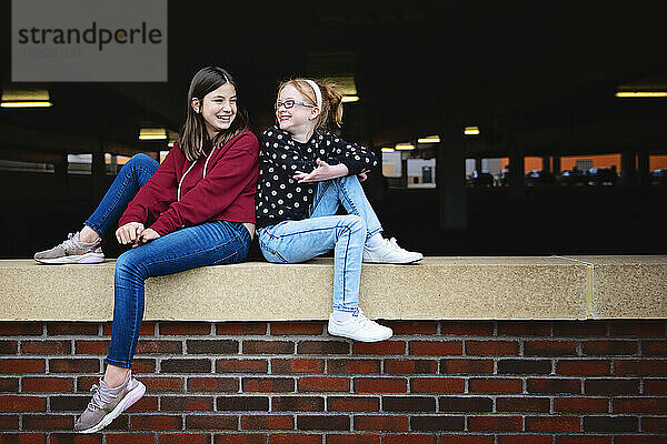 Zwei glückliche Mädchen zwischen zwei Jahren sitzen auf einer Mauer und unterhalten sich.