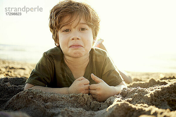 Porträt eines niedlichen Jungen  der an einem sonnigen Tag am Strand im Sand liegt