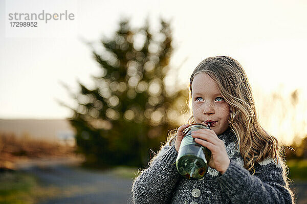 Süßes Mädchen nippt an einem frischen Getränk aus einem Glas und schaut nach oben  während sie sich im Park bei Sonnenuntergang ausruht
