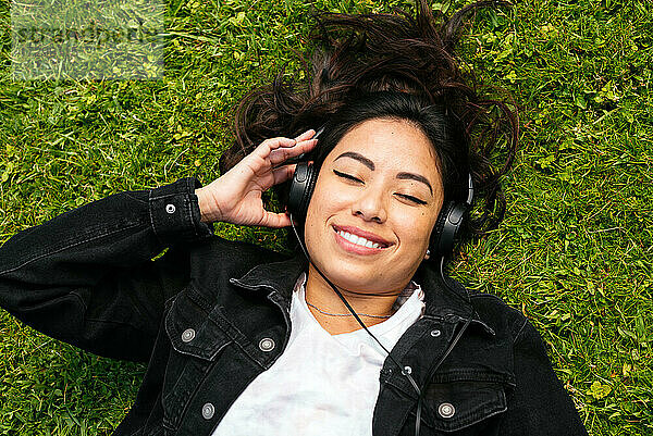 Draufsicht auf eine fröhliche Frau  die im Gras liegt und Musik hört