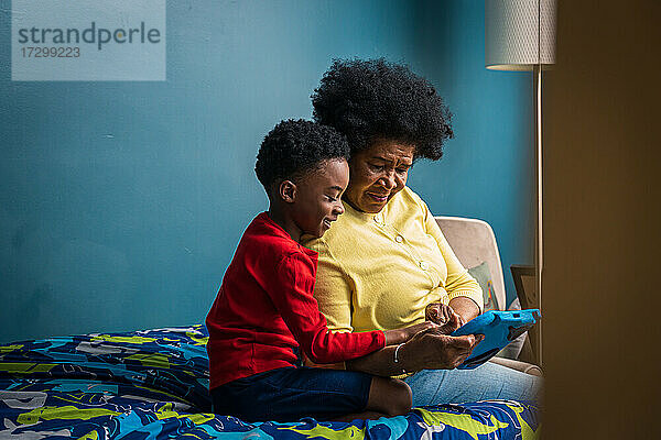 Lächelnder Junge und Großmutter benutzen ein digitales Tablet  während sie zu Hause im Schlafzimmer sitzen