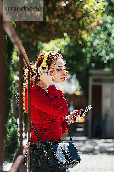 Junge rothaarige Frau entspannt sich  während sie mit ihrem Handy und gelben Kopfhörern in einem städtischen Raum Musik hört. Bekleidet mit einer roten Bluse.