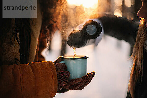 Frau gießt Tee für Freundin in Tasse stehend im Park im Winter