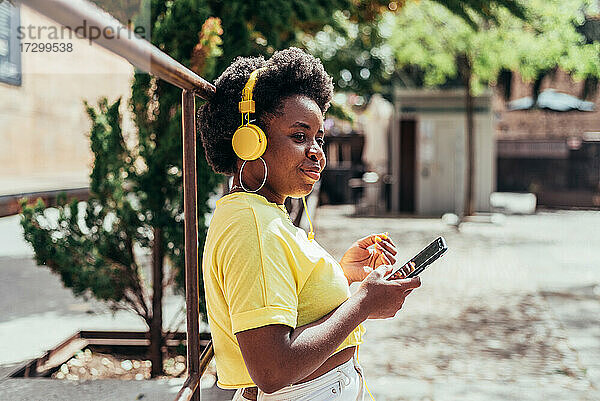 Seitenansicht eines schwarzen Mädchens mit Afro-Haar und Ohrringen  das mit ihrem Handy und gelben Kopfhörern in einem städtischen Raum Musik hört.