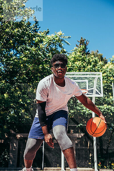 Schwarzer afroamerikanischer Junge mit Sonnenbrille spielt Basketball auf einem städtischen Platz.