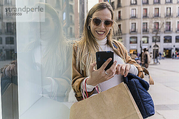 Fröhliche Frau beim Einkaufen in der Stadt. Sie benutzt ihr Smartphone.