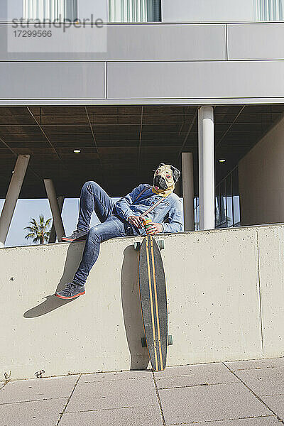 Mann mit Mopsmaske  der auf einer Mauer liegt und ein Longboard trägt.