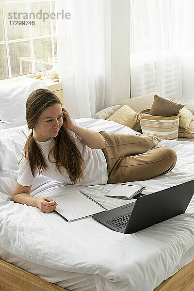 Frau im weißen T-Shirt liegt auf dem Bett und schaut auf den Laptop