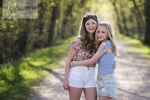 Zwei hübsche Teenager-Mädchen  beste Freundinnen  stehen auf einer von Bäumen gesäumten Straße.